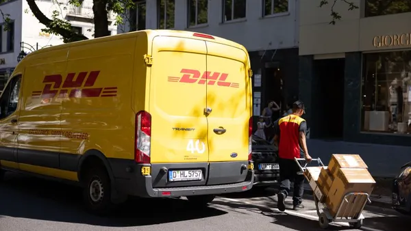 A person in a DHL uniform moves a dolly with packages on a street. A DHL delivery van is parked on the street.