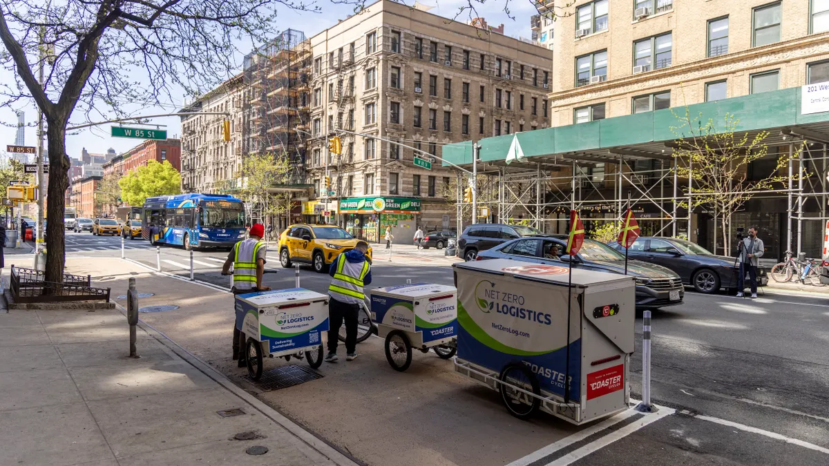 Workers wearing reflective vests pull hand carts with large boxes on the side of a city street.