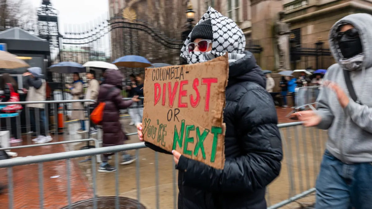 A protester outside the gates of Columbia University holds a sign saying, in all capital letters, "Columbia - divest or you're next."